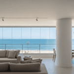 sala de estar de alto padrão em um apartamento na orla de Salvador. Através de uma grande janela panorâmica de vidro, vê-se a praia do Rio Vermelho sob um dia ensolarado. As paredes internas do apartamento estão perfeitamente lisas e pintadas em um tom off-white fosco, sem nenhuma imperfeição.