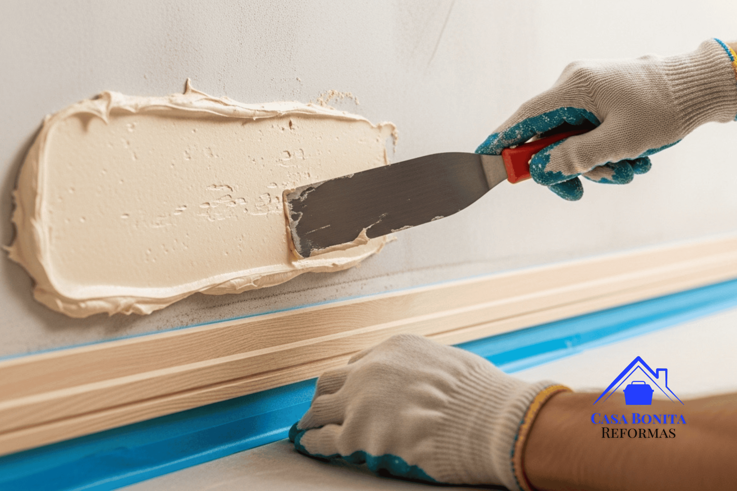 A Correção Técnica (O Fim do Erro PVA) pintor profissional usando luvas, segurando uma espátula de aço larga. Ele está aplicando uma camada espessa e firme de massa acrílica sobre uma parede que foi previamente raspada até o reboco áspero. O rodapé de madeira nobre logo abaixo está perfeitamente protegido com fita adesiva azul de precisão.
