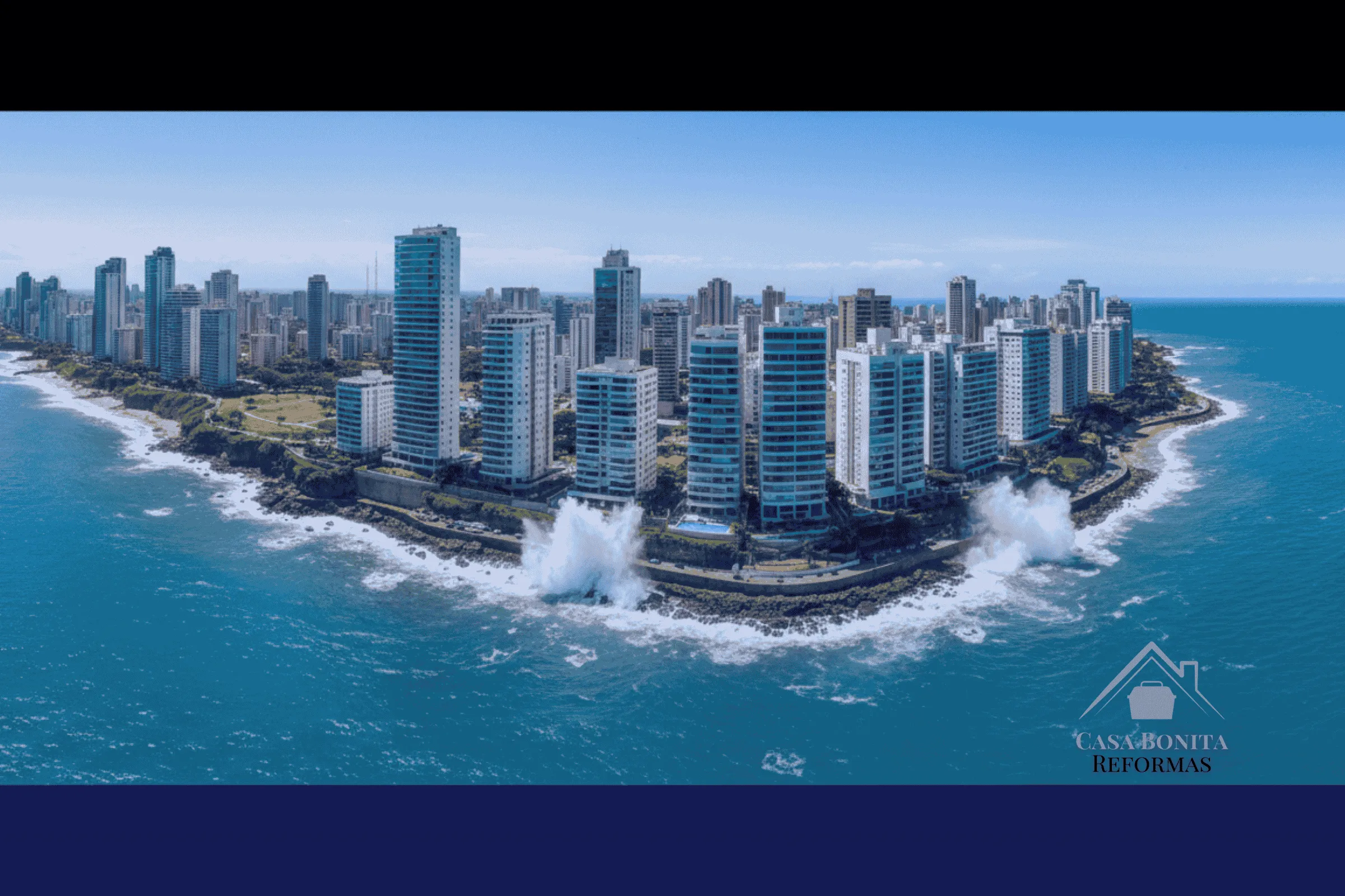 O 'Mapa da Maresia' de Salvador Especificação Técnica por Bairro Vista aérea panorâmica e ensolarada da região do Corredor da Vitória ou Barra em Salvador. A imagem mostra edifícios residenciais altos e luxuosos construídos extremamente próximos à beira do mar azul-turquesa. As ondas batem nas pedras próximas aos prédios, transmitindo a ideia da maresia constante atingindo as fachadas. O céu é azul claro. Estilo fotografia de paisagem urbana.