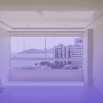 sala de estar em um apartamento de alto padrão em Salvador, recém-pintada em um tom off-white sofisticado. Através de uma grande janela panorâmica, vê-se a vista deslumbrante da Baía de Todos os Santos e edifícios na orla sob sol forte. A sala está impecável, sem móveis, destacando apenas a qualidade perfeita do acabamento das paredes e do teto. A luz natural do sol entra pela janela, valorizando a textura da tinta.