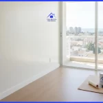 paredes recém-pintadas em um tom off-white aveludado perfeito refletem a luz natural que entra por uma grande janela (com uma sutil vista urbana de Salvador ao fundo). Em um canto da sala, sobre um papelão de proteção perfeitamente alinhado no chão, descansam um rolo de pintura premium limpo e uma lata de tinta fechada.