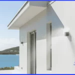 fachada de uma casa de alto padrão contemporânea, com paredes perfeitamente lisas e pintadas de branco ofuscante. A casa está localizada à beira-mar, com a Baía de Todos os Santos ao fundo. O sol forte (radiação UV) bate diretamente na pintura, enquanto uma leve névoa visível vinda do mar (maresia) atinge a fachada.