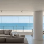 sala de estar de alto padrão em um apartamento na orla de Salvador. Através de uma grande janela panorâmica de vidro, vê-se a praia do Rio Vermelho sob um dia ensolarado. As paredes internas do apartamento estão perfeitamente lisas e pintadas em um tom off-white fosco, sem nenhuma imperfeição.